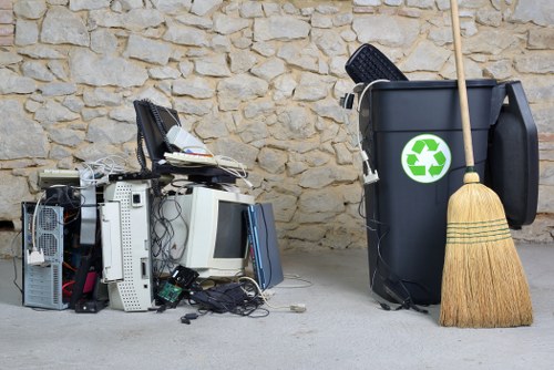 A pile of discarded electronic waste including several computer towers, monitors, keyboards, and miscellaneous electronic components is placed on a light-colored pavement in front of a textured beige brick wall. The electronic items are tangled with cables and wires, with some components leaning against each other, creating a disorganized stack. Next to the pile, there is a large black recycling bin with a green recycling symbol, partially filled with waste items including a black plastic container and a wooden-handled broom resting against it. The scene appears to be outdoors in an urban or residential environment, possibly on a driveway or back alley, highlighting waste collection or disposal as part of rubbish removal services in Lambeth or nearby areas. The setting is well-lit, with natural daylight illuminating the scene clearly.