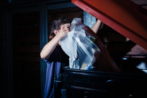 A person wearing a dark shirt is seen disposing of a clear plastic bag filled with various pieces of rubbish into a large black waste container outdoors. The container is positioned in front of a wooden building with horizontal planks, and there are blue metal support beams visible in the background. The scene is captured in natural daylight, highlighting the texture of the plastic bag and the surrounding environment. This image illustrates the process of rubbish removal typical of services provided by Office Clearance Lambeth, relevant to waste management and clearance tasks within the Lambeth area, which is located in South London with postcode postcode inferred from the Lambeth region.