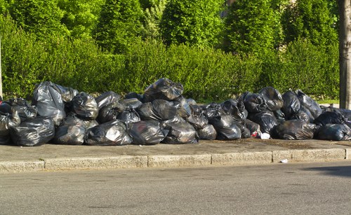 A large accumulation of black rubbish bags, filled and tied, piled along the edge of a paved street or driveway, with a curb separating them from a green, leafy background composed of trees and dense shrubbery. The bags appear to be made of plastic with a shiny surface, and they are stacked in a somewhat uneven manner, suggesting a recent or ongoing rubbish removal process. The setting suggests an outdoor area possibly near a residential or commercial property in Lambeth, within the postcode area, where Office Clearance Lambeth provides waste disposal services. The natural lighting highlights the texture of the plastic bags and the contrast with the lush greenery behind them, emphasizing the urban waste collection scene in a suburban or urban environment typical of London boroughs. This image visually supports rubbish removal services by illustrating a typical collection or disposal scene.