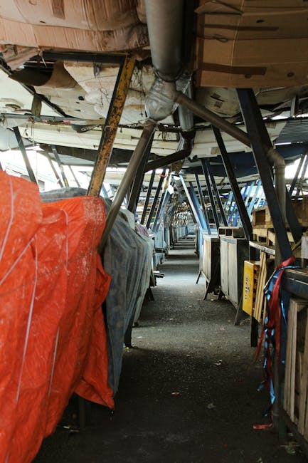 The image depicts the interior of a boat or aircraft with a narrow, enclosed corridor running through the middle. On the left side, there are several large objects covered with protective tarps in sha