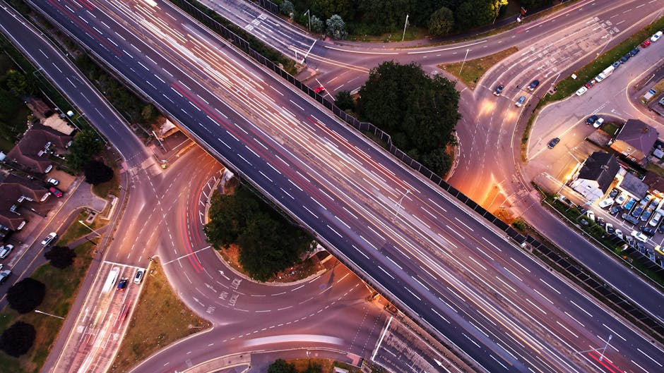 An aerial view of a multi-lane urban highway interchange during dusk, capturing light trails from moving vehicles that create streaks of white and red across the dark asphalt surface. The interchange 