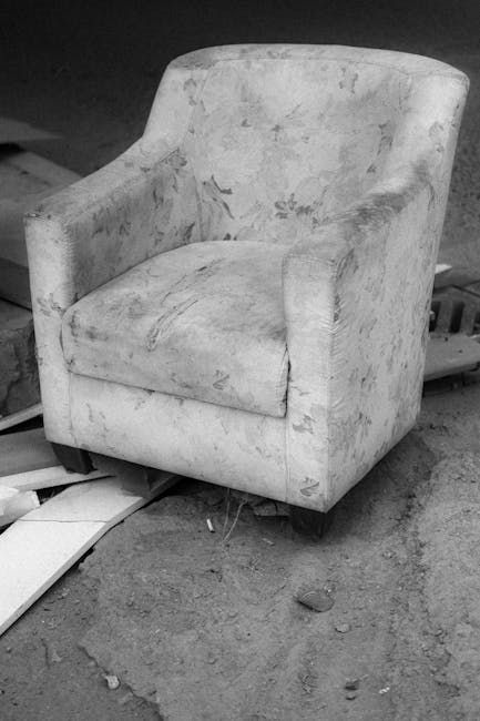 Image showing two vintage upholstered armchairs placed on a concrete or wooden floor in an indoor setting, possibly a storage or clearance area. The armchair on the left features a tall, straight back