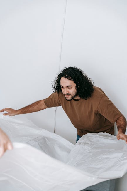 A person with curly brown hair, wearing a green t-shirt and blue jeans, is bent over and carefully removing a large white mattress from a bed in what appears to be a clean, well-lit interior space. Th