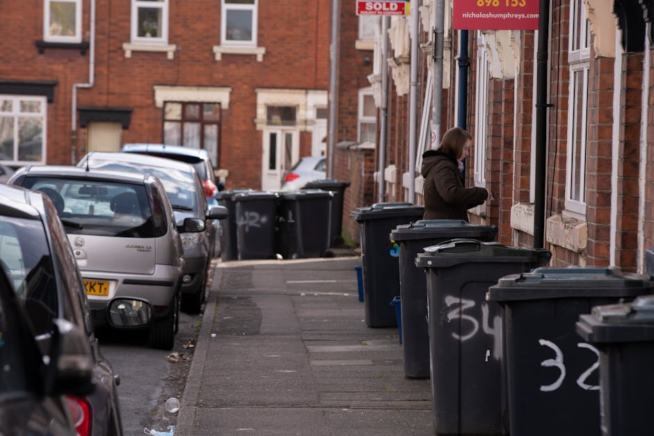 A narrow urban street scene featuring multiple black wheelie bins placed along the pavement, with some lids slightly open and marked with white graffiti. To the right, a woman dressed in a dark jacket