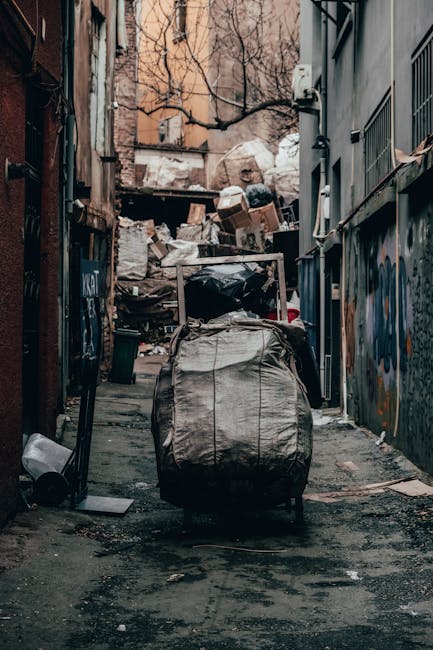 A narrow urban street scene featuring multiple black wheelie bins placed along the pavement, with some lids slightly open and marked with white graffiti. To the right, a woman dressed in a dark jacket