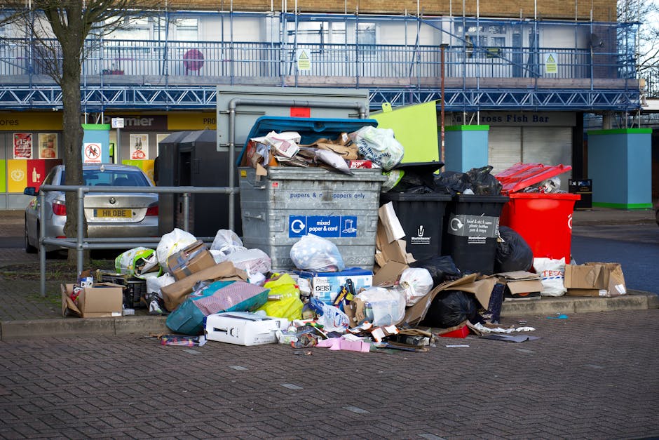 A collection of discarded household items and waste materials stacked on a sidewalk beside a building. The pile includes a partially folded and worn Persian-style rug with intricate pink, purple, and