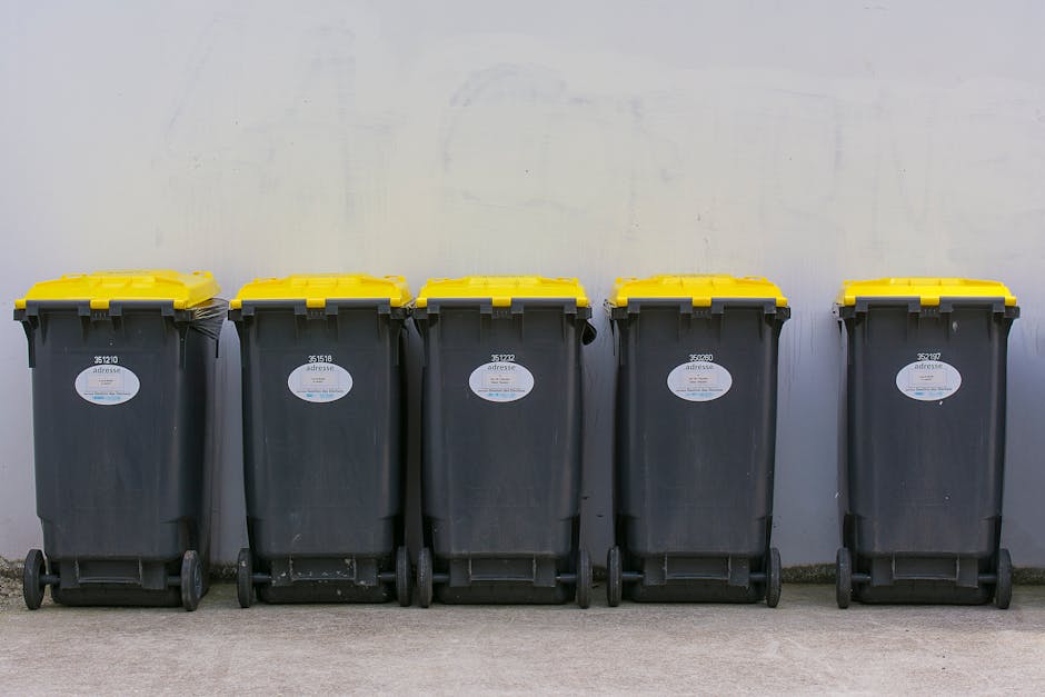A man with dark hair, wearing a black t-shirt with white graphic text, is seen disposing of a large white plastic bag filled with rubbish into a cylindrical metal trash bin. The trash bin has a shiny,